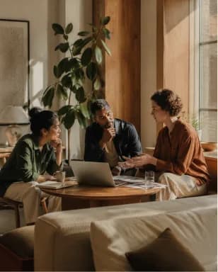 Three people talking around laptops in a plant-filled living room.